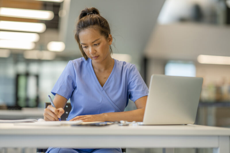A nurse in scrubs sits in a hospital environment in front of a laptop computer and is writing notes on a notepad.
